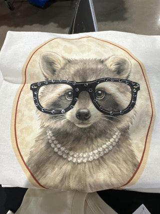 Tee with a design of a raccoon face wearing glasses and a pearl necklace on an oatmeal background